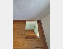 Blick von unten auf eine schmale Holztreppe, die durch einen rechteckigen Durchgang nach oben führt. Die Wände und Verkleidungen sind aus Holzpaneelen, darüber eine weiße, strukturierte Decke und angrenzende weiße Wandflächen.