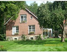 Ziegelsteinhaus mit Satteldach und weiß gerahmten Fenstern, umgeben von einem gepflegten Garten mit Rasenfläche und Obstbäumen. Im Hintergrund sind ein Wintergartenanbau sowie weitere Pflanzen und Bäume sichtbar.