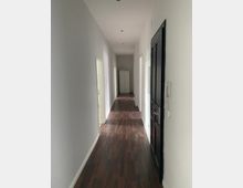 Langer, schmaler Flur in einem Wohngebäude mit weißen Wänden und dunklem Holzboden. Türen befinden sich an beiden Seiten, und am Ende des Flurs steht ein weißer Schrank.