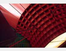 Decke eines Innenraums mit roten, wabenförmigen Metallstrukturen und einem gebogenen gelben Lichtpanel; im Hintergrund sind grüne Wandfliesen erkennbar.