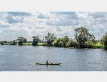 Ein Fluss mit ruhiger Wasseroberfläche, umgeben von Bäumen und Wiesen im Hintergrund. Im Vordergrund paddelt eine Person in einem gelben Kajak unter einem bewölkten Himmel.