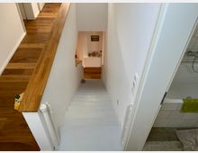 Blick auf eine weiße Treppe, die von einem oberen Stockwerk mit Holzfußboden hinunterführt. Links befindet sich ein hölzernes Geländer, rechts ist ein Badezimmer mit Badewanne und Fliesenboden zu sehen.