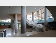 Innenansicht eines modernen öffentlichen Gebäudes mit großen Fenstern, durch die Tageslicht hereinfällt. Zu sehen sind eine breite Treppe mit Metallgeländern, große runde Betonpfeiler und glatte Oberflächen mit Holz- und Betonakzenten.