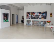 Galerieraum mit weißen Wänden und hellbeigem Boden, an denen großformatige Fotografien hängen, darunter Bilder eines Cockpits. Auf einem weißen Tisch rechts steht eine Vase mit bunten Blumen. Im Hintergrund sind Türen und weitere Kunstwerke sichtbar.