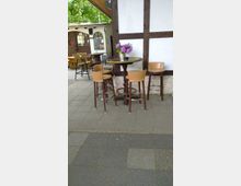 Außenbereich eines Cafés mit einem runden Stehtisch aus Holz, umgeben von vier Barhockern. Im Hintergrund sind ein Fachwerkhaus, Holztische und -stühle sowie grüne Bäume zu sehen.