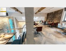 Ein modernes Restaurant mit Backsteinwänden, einem hölzernen Fischgrätenboden und freiliegenden Holzbalken. Tische sind mit Gläsern, Tellern und Stoffservietten gedeckt, während im Hintergrund ein leuchtendes Neonschild mit der Aufschrift "Oderberger" sichtbar ist.