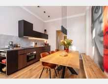 Moderne Wohnküche mit dunkler, griffsloser Einbauzeile und grauer Rückwand, Holzboden sowie langem Holzesstisch mit Lederstühlen. Über dem Tisch hängen schwarze Pendelleuchten; an der rechten Wand befindet sich ein großformatiges Wandbild.