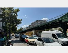 Straßenszene in einer Stadt mit mehreren Autos im Verkehr, einem Bürgersteig und Fahrrädern auf der linken Seite. Eine grüne Hochbahnbrücke verläuft über die Straße, flankiert von Bäumen und Gebäuden im Hintergrund.