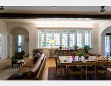 Wohnzimmer und Esszimmer in einem hellen, historischen Stil mit Holzboden. Der Raum verfügt über große Sprossenfenster, die viel Licht hereinlassen, einen braunen Ledersessel, einen Esstisch mit Holzstühlen und dekorative Pflanzen auf einem Heizkörper vor den Fenstern.