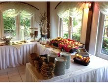 Ein Frühstücksbuffet in einem hellen Raum mit großen Fenstern, durch die grüner Garten sichtbar ist. Auf den mit weißen Tischdecken bedeckten Tischen stehen Brotkörbe, Tellerstapel, Müslispender, Obst und weitere Frühstückszutaten.