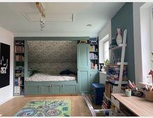 Kinderzimmer mit eingebautem Bett in einer Nische, flankiert von Regalen mit Büchern und Spielzeug. Rechts stehen ein Schreibtisch und Regale mit Brettspielen, während ein bunter Spielteppich auf dem Holzboden liegt.