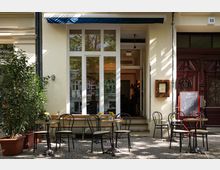 Fassade eines Cafés mit heller Wand, großen verglasten Türen und einer blauen Markise. Davor stehen kleine runde Tische mit schwarzen Stühlen auf einem gepflasterten Gehweg, flankiert von Pflanzkübeln; eine Kreidetafel mit „Welcome!”-Schriftzug steht an der Seite.