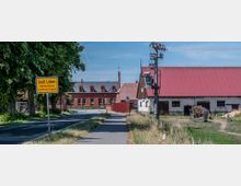 Ortseingang Groß Lüben im Landkreis Prignitz, mit gelbem Ortsschild am Straßenrand, einem Gehweg daneben, einem roten Ziegelgebäude im Hintergrund und einer Scheune mit rotem Dach auf der rechten Seite. Im Vordergrund befinden sich Strommasten und Grünflächen.