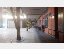 Weitläufiger Innenraum einer Industriehalle mit hohen Decken, Metallträgern und orangefarbenen Wänden auf der rechten Seite. Die Halle ist in Abschnitte unterteilt, mit gelb-schwarz markierten Stützpfeilern und einer Person in Sicherheitskleidung im Hintergrund.