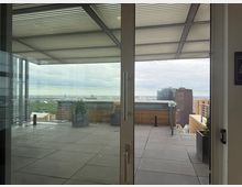 View through large glass doors onto a modern rooftop terrace with gray tiled flooring, metal railings, and square planters under a slatted canopy. A city skyline with several high-rise buildings is visible in the distance beyond the terrace edge.