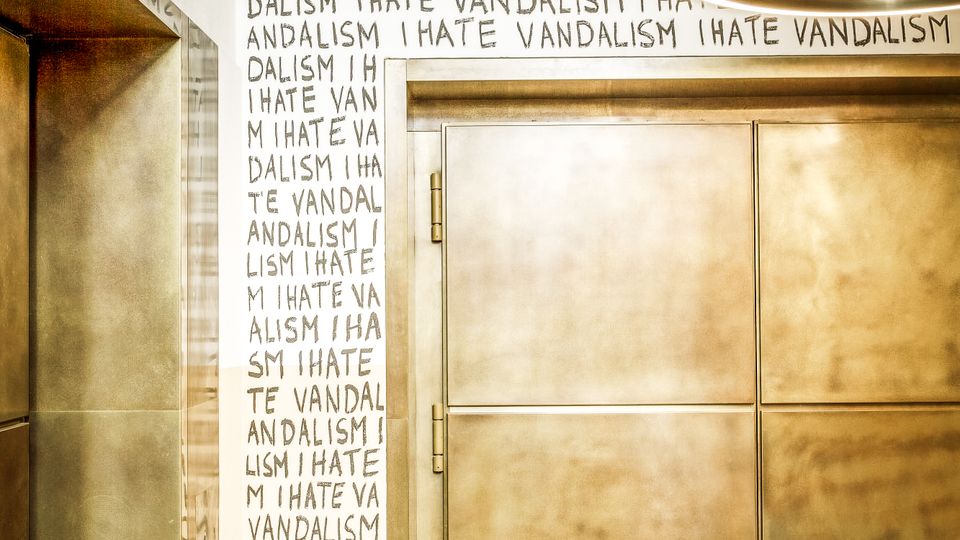 Innenansicht eines Flurs oder Eingangsbereichs mit großen, metallischen Doppeltüren und einer runden Deckenleuchte. Die Wand ist über mehrere Reihen mit dem wiederholten Schriftzug „I HATE VANDALISM“ in schwarzer Handschrift bedeckt.