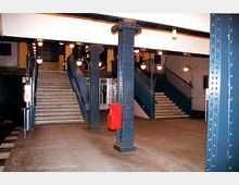 Eingangsbereich einer U-Bahn-Station mit Treppen, die nach oben führen, blau gestrichenen Stahlpfeilern und historischen Laternen. Eine rote Notruf- oder Feuerlöscherbox ist an einem der Pfeiler angebracht.