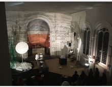 Eine Innenaufnahme eines historischen Kirchenraums, der für eine künstlerische oder multimediale Inszenierung genutzt wird. Große, gewölbte Fenster mit Bleiverglasung und projizierte Texte bedecken die Wände und den Altarbereich, während mehrere Personen in weißen Gewändern mit Lichtquellen und Requisiten auf einer Bühne agieren.
