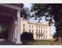 Gelbes Herrenhaus im klassizistischen Stil mit symmetrischer Fassade, verzierten Fenstern und einem Balkon, umgeben von Bäumen und einer Rasenfläche. Im Vordergrund ein Säulengang, der teilweise den Blick auf das Gebäude einrahmt.