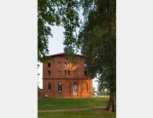 Backsteingebäude mit hohen Rundbogenfenstern, umgeben von grüner Wiese und schattenspendenden Bäumen; der Himmel ist klar und lichtdurchflutet.