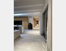 Moderner Wohnbereich mit hellen Fliesenboden, einer offenen Küche mit Holzfronten und Marmorarbeitsfläche sowie einer großen Glasfront mit Vorhängen. Im Hintergrund führt ein langer, beleuchteter Flur zu einer Wand mit einem Bild.