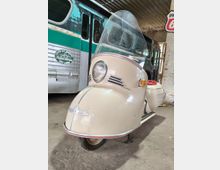 Ein cremefarbener, retro Motorroller mit großer Windschutzscheibe steht in einer überdachten Halle mit Betonboden. Im Hintergrund sind ein grün-silberner Linienbus und eine Backsteinwand mit einem Route-66-Schild zu sehen.