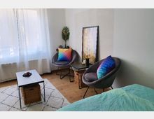 Wohnzimmer mit hellen Wänden und Holzboden, ausgestattet mit zwei grauen Sesseln, die je ein buntes Kissen tragen. Ein niedriger Couchtisch mit Rattan-Korb darunter sowie ein Teppich mit geometrischem Muster und ein großes Fenster mit weißen Vorhängen bestimmen den Raum.