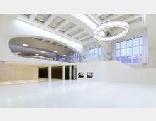 Großer, moderner Veranstaltungsraum mit weißem Boden, hohen Decken und einer Galerieebene mit Glasgeländer. Ein auffälliger, runder Kronleuchter hängt in der Mitte, und große Fenster lassen Tageslicht herein; dekorative Deckenpaneele ergänzen das minimalistische Design.