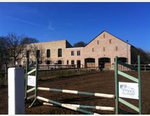 Ein ländlicher Reitplatz mit einer niedrigen Hindernisstange im Vordergrund, umgeben von einem Holzzaun. Im Hintergrund stehen Gebäude aus hellbraunem Backstein mit großen Torbögen und Fenstern, die in eine bäuerliche oder reitsportliche Anlage eingebettet sind.