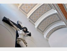 Kreuzigungsdarstellung in einer Kirche mit einer realistischen Jesusfigur am schwarzen Kreuz; im Hintergrund sind weiße, gewölbte Decken mit geometrischen, farbig verzierten Mustern zu sehen.