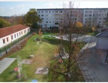 Wohnsiedlung mit einem zentralen Grünbereich. Links ein einstöckiges Gebäude mit roten Fensterläden, dahinter ein mehrstöckiges Wohnhaus; Gehwege und Bäume sind im Gelände verteilt.