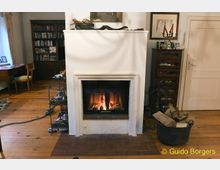 Ein gemütliches Wohnzimmer mit einem Kamin aus weißem Putz, in dem ein Feuer brennt. Im Raum befinden sich Holzmöbel, darunter ein Schreibtisch und ein Bücherregal, sowie ein Holzkorb neben dem Kamin.