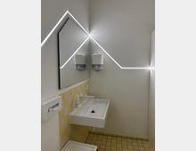 Moderne, minimalistische Toilette mit weißem Waschbecken, gelb-weiß gefliester Wand- und Bodenfläche und einem großen Spiegel. Auffällig sind die diagonalen LED-Lichtlinien an der Wand, die ein geometrisches Design betonen.