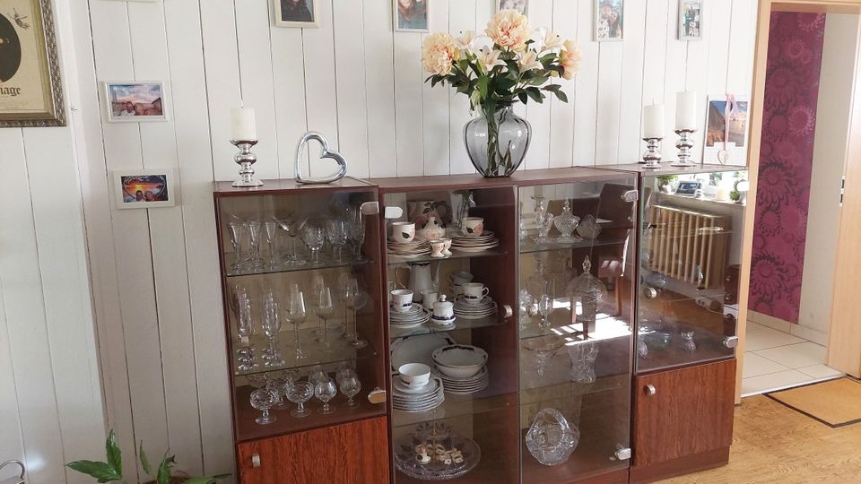 Wohnzimmerbereich mit einer langen Glasvitrine aus Holz, gefüllt mit Gläsern und Porzellan, vor einer weiß vertäfelten Wand mit mehreren gerahmten Fotos. Auf der Vitrine stehen Kerzenhalter, ein Herzdeko-Element und eine große Vase mit Blumen; rechts führt eine offene Tür in einen Flur mit gemusterter Tapete.