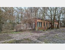 Verlassenes, niedriges Backsteingebäude mit flachem Dach und einer Reihe großer Fenster an der Seitenfassade, teilweise von Büschen und Bäumen überwuchert. Davor liegt ein verwitterter, moosiger Asphalt- oder Betonplatz mit Laub, umgeben von dichtem, kahlem Wald.