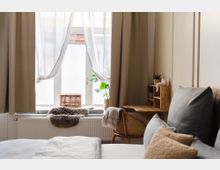 Gemütliches Schlafzimmer mit hellem Bett und blauen sowie beigen Kissen. Das Zimmer hat ein Fenster mit beigefarbenen Vorhängen, eine Fensterbank mit Dekorationen wie einem Fell, einem Korb und einer Pflanze, sowie einen Holzstuhl und ein Wandregal aus Holz.