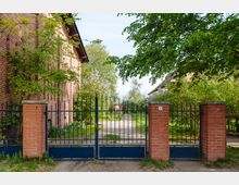 Ein Eingangstor aus schwarzem Metall mit roten Ziegelpfeilern führt zu einem schmalen Weg, der durch einen grünen Garten mit Bäumen und Sträuchern verläuft. Links und rechts des Tores befinden sich Backsteingebäude mit Satteldächern.