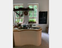 Helle Küche mit geschwungener Kochinsel aus Holz und Edelstahlspüle im Vordergrund. Im Hintergrund drei Fenster mit Blick auf grüne Bäume, dekoriert mit Kräutertöpfen und rustikalen Zweigen, sowie eine mit Kreide bemalte Tafel.