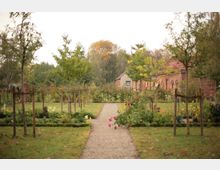 Ein gepflegter Garten mit einem Kiesweg, der zwischen Blumenbeeten und jungen Bäumen verläuft. Im Hintergrund stehen Backsteingebäude und ein Wohnhaus, umgeben von Bäumen und Sträuchern.