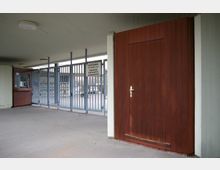 Überdachter Eingangsbereich mit einer roten Holztür auf der rechten Seite und einer blauen Metallgitterabsperrung, die den Zugang begrenzt; ein Hinweisschild mit Regeln für den Durchgang ist an der Gitterabsperrung angebracht.