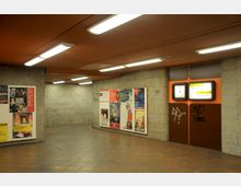 Unterirdischer U-Bahn-Bereich mit gefliesten Wänden und Boden in warmen Tönen. An einer Wand befinden sich Plakate, an einer anderen eine stationäre Anzeige mit einer Uhr und Wegweisern zu den Ausgängen.