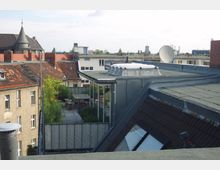 Blick über die Dächer einer Stadt mit Ziegel- und Flachdächern, einer begrünten Dachterrasse mit Sitzmöbeln und hinterliegenden Wohnhäusern. Im Hintergrund sind ein Turm und weitere moderne Gebäude zu sehen.