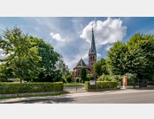 Roter Backstein-Kirchbau mit hohem Spitzdach-Turm, umgeben von Bäumen und einem gepflegten Garten mit Zaun; im Vordergrund eine Straße mit einem Bürgersteig.