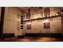 Industrieller Innenraum mit hohen, rauen Wänden aus verputztem Backstein, kleinen verschlossenen Fenstern und einer hängenden Lichtinstallation. Im unteren Bereich befinden sich eine Bank mit dunklen Polstern und ein Holzboden.