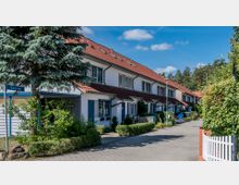 Reihenhaussiedlung mit weiß gestrichenen Häusern, roten Ziegeldächern und blauen Fensterläden. Vor den Häusern befinden sich Vorgärten mit Pflanzen und ein gepflasterter Gehweg, umgeben von Bäumen und Sträuchern.