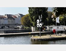 Hafenbereich mit Wasserweg, umgeben von Bäumen und Gebäuden im Hintergrund. Im Vordergrund sind ein Steg, Signalleuchten auf einem Mast und ein Teil eines Anlegers zu sehen, hinter dem kleinere Boote und eine Brücke erkennbar sind.