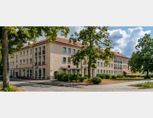 Wohngebäude mit heller Fassade und rotem Ziegeldach, umgeben von Bäumen und einer gepflasterten Gehwegfläche. Im Vordergrund sind parkende Fahrräder, ein Verkehrsschild und gepflegte Grünflächen zu sehen.