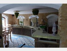 Innenansicht eines Restaurants mit rustikalem Design: Wände aus sichtbarem Ziegelstein, eine verzierte Holztheke mit Mosaikfliesen und ein gefliester Boden mit geometrischen Mustern. Im Hintergrund sind große Fensterbögen, Dekorationen und Pflanzen sichtbar.