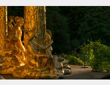 Goldene Statuengruppe vor kunstvoll verzierten Säulen, umgeben von dichtem Grün und beleuchtet von warmem Licht. Details zeigen asiatisch inspirierte Figuren in sitzenden und stehenden Posen.