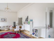 Helles Wohnzimmer mit einem beigefarbenen Ecksofa, Holzboden und einem großen, roten Orientteppich. Im Raum befinden sich ein Esstisch mit weißen Stühlen, mehrere weiße Regalwürfel, eine Tür mit Glaseinsatz und eine goldene Deckenlampe.